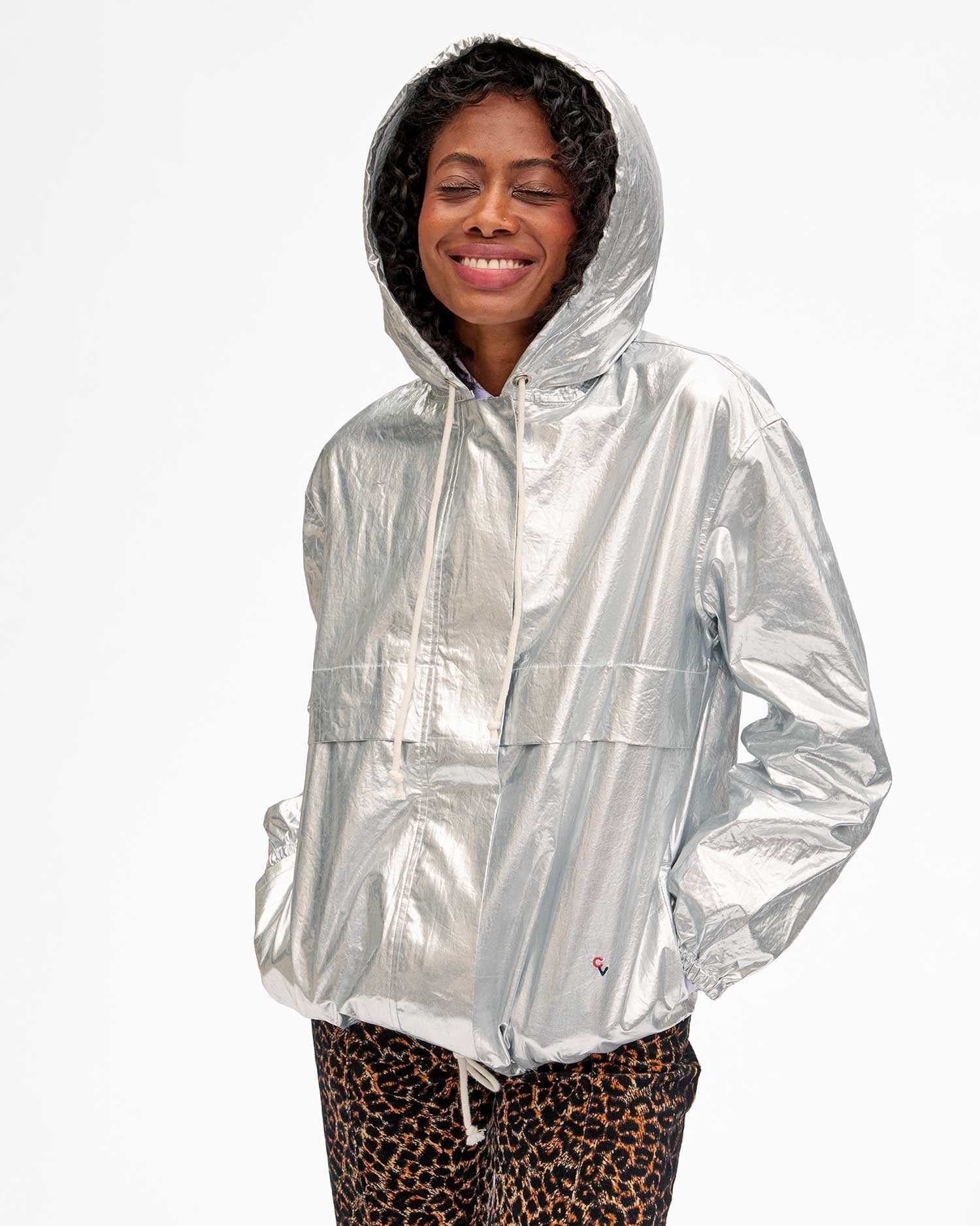 A person with short curly hair smiles with closed eyes, wearing the Theo Anorak Jacket by Clare Vivier. The jacket is shiny silver and features elastic sleeves, and they pair it with leopard print pants. With hands in pockets, they stand against a plain white background.