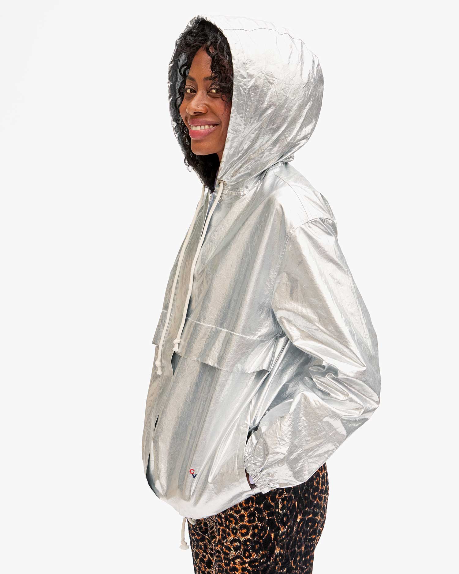 A person wearing the Clare Vivier Theo Anorak Jacket, complete with a metallic coating and drawstring hood, smiles while standing sideways. The jacket also showcases a front pocket. They pair it with leopard print pants against a plain white background.