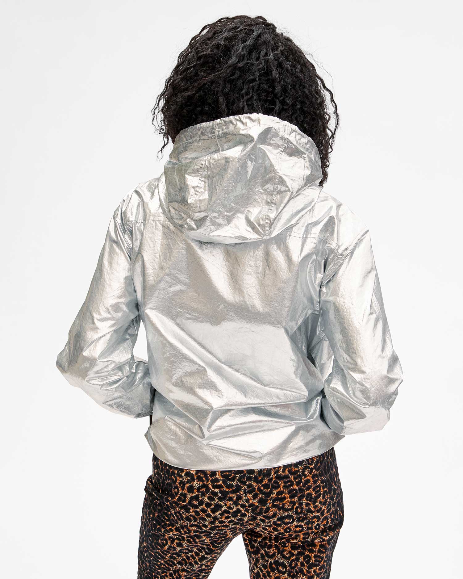 A person with curly hair is wearing the Clare Vivier Theo Anorak Jacket, a shiny silver hooded jacket with side pockets, paired with leopard print pants, standing facing away from the camera against a plain background.