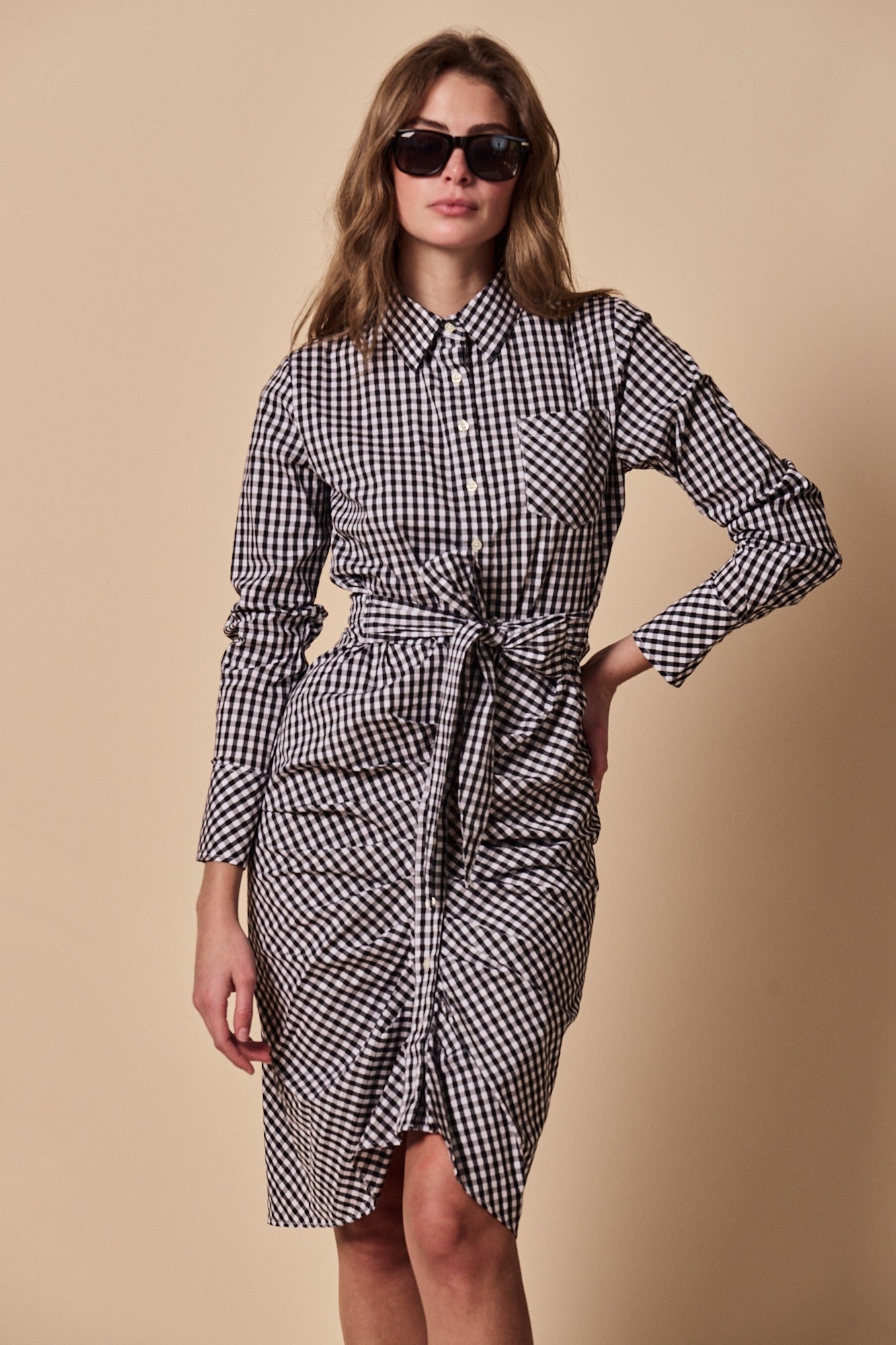 A woman wearing THE SHIRT's The Wrap Front Dress—a black-and-white checkered, long sleeve shirt dress with a waist tie—stands against a beige background.