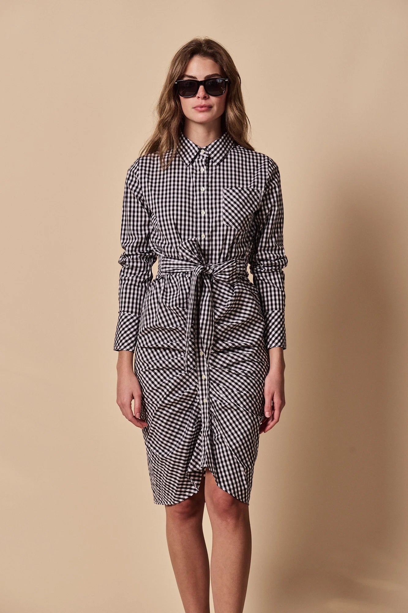 A woman wearing THE SHIRT’s The Wrap Front Dress, a black-and-white checkered long sleeve shirt dress with a waist tie, stands in front of a beige background.