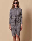 A woman wearing THE SHIRT’s The Wrap Front Dress, a black-and-white checkered long sleeve shirt dress with a waist tie, stands in front of a beige background.