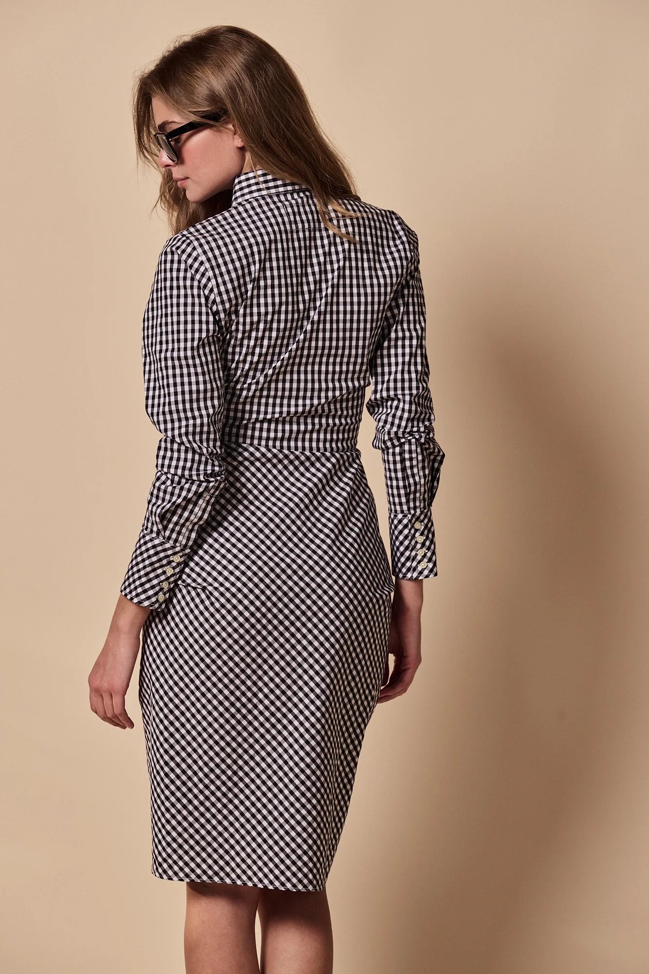 A woman wearing sunglasses and The Wrap Front Dress by THE SHIRT, featuring black-and-white checks and long sleeves, stands with her back to the camera against a plain beige background.