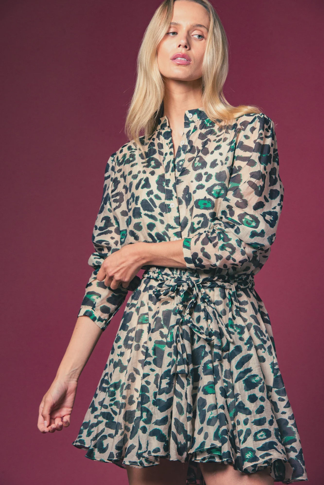 A woman poses against a maroon background, wearing THE SHIRT's Jenica Dress with No Gape button technology, featuring a green and black leopard print and a cinched, flared skirt.
