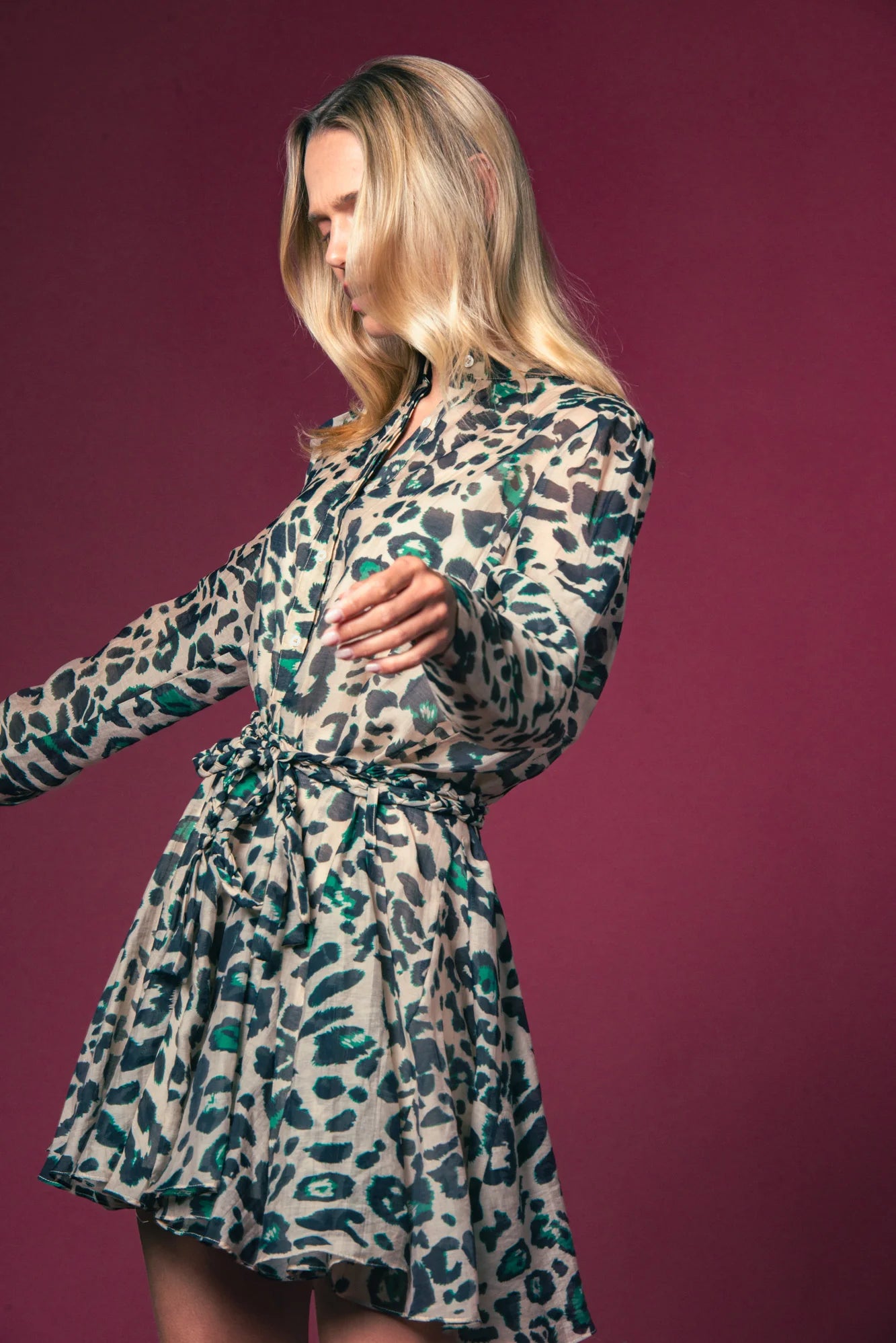A woman with long blonde hair models the Jenica Dress by THE SHIRT, a patterned shirtdress with green and black spots and a braided rope belt, standing against a solid maroon background.