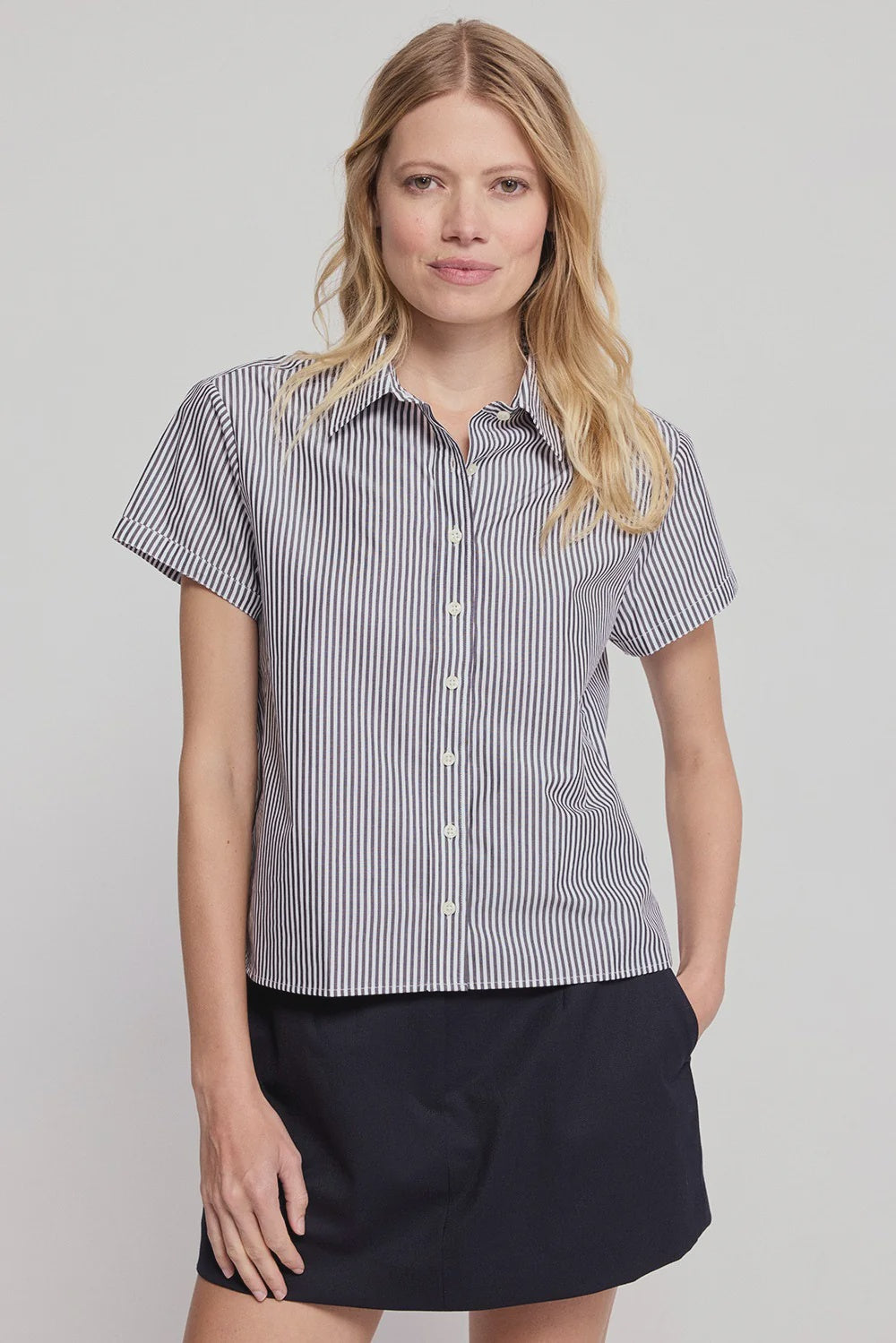 A woman with long blonde hair wears THE SHIRT’s Boxy SS Cropped Shirt—a black-and-white vertically striped button-up—paired with a black skirt, standing with one hand in her pocket against a light gray background.