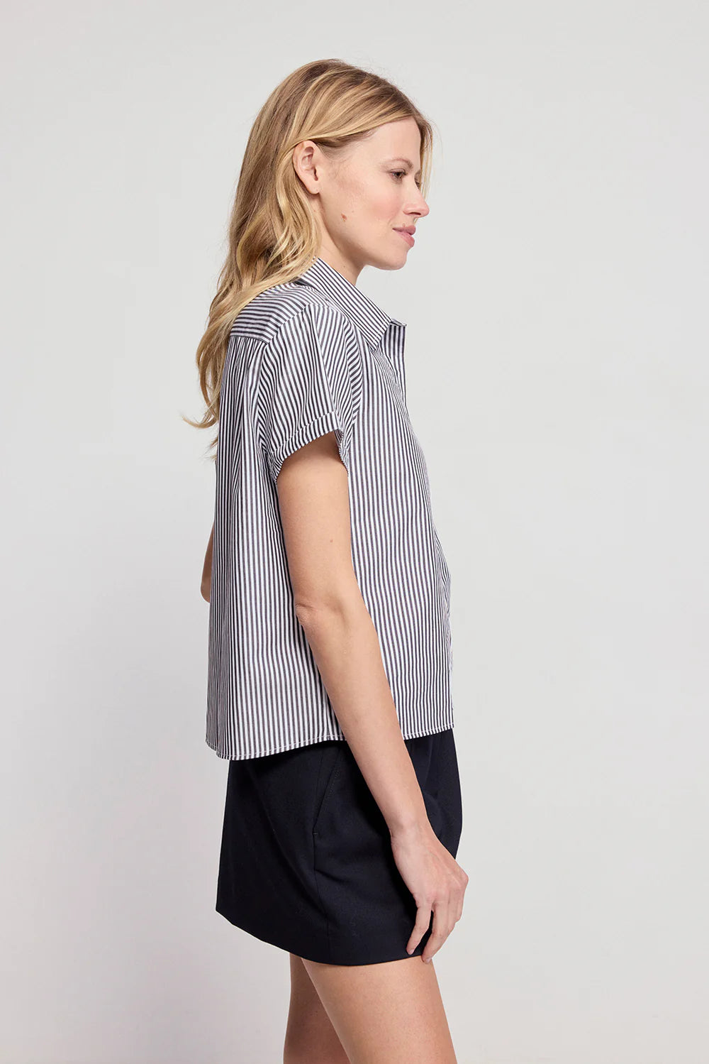 A blonde woman wears THE SHIRT's Boxy SS Cropped Shirt in black-and-white stripes with a dark skirt. The untucked, short-sleeved, boxy top stands out as she poses in profile against a light background.