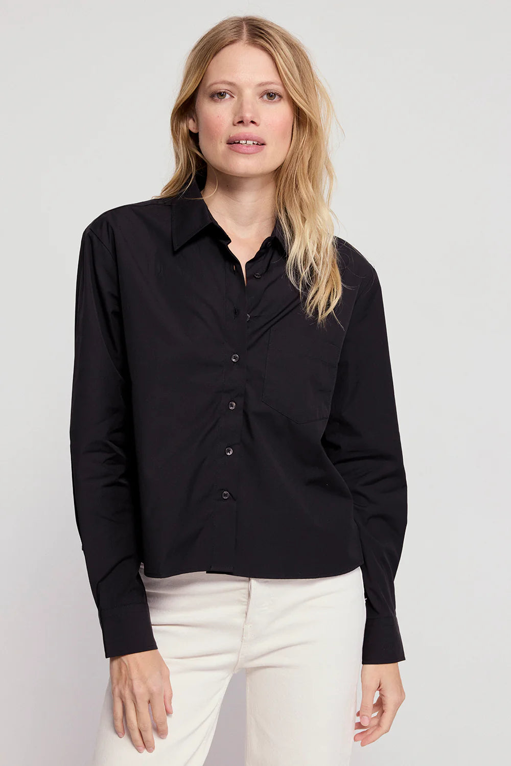 A woman with long blonde hair wears THE SHIRT's The LS Boxy Crop, a black button-up, and white pants, standing against a plain light gray background.