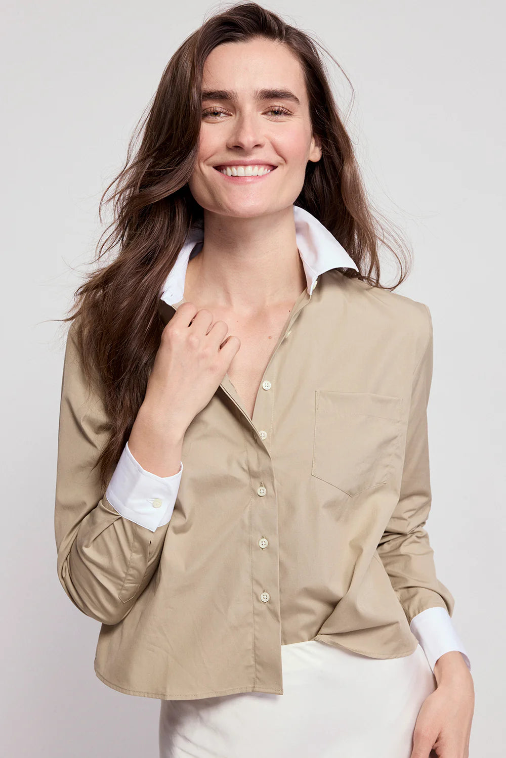 A woman with long brown hair wears THE SHIRT’s The LS Boxy Crop beige button-up, featuring No Gape button technology. She smiles confidently, holding the lapel against a plain, light background.