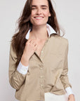 A woman with long brown hair wears THE SHIRT’s The LS Boxy Crop beige button-up, featuring No Gape button technology. She smiles confidently, holding the lapel against a plain, light background.