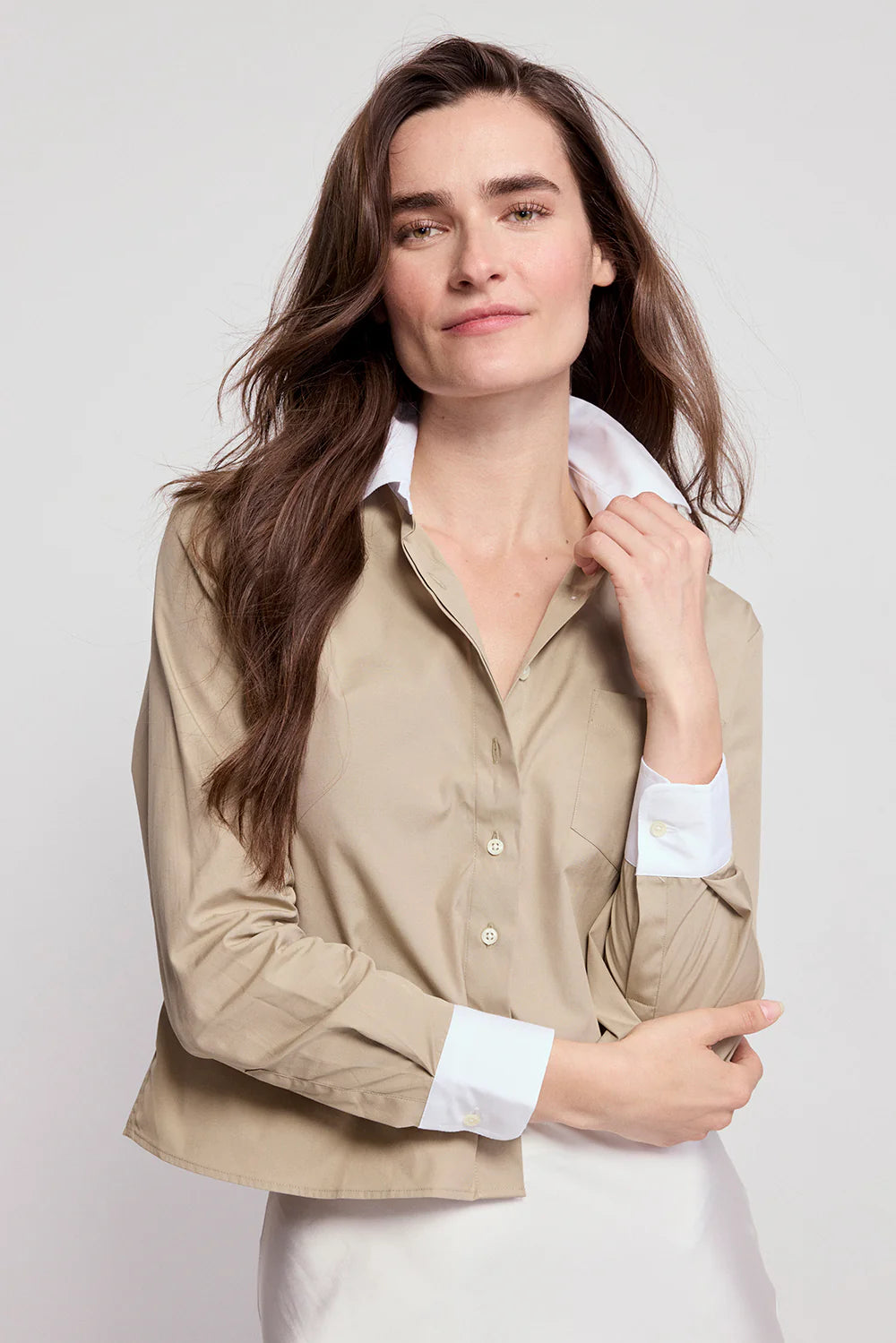 A woman with long brown hair wears The LS Boxy Crop by THE SHIRT, a beige button-up with white cuffs and collar featuring No Gape technology, standing against a plain light background and smiling softly.