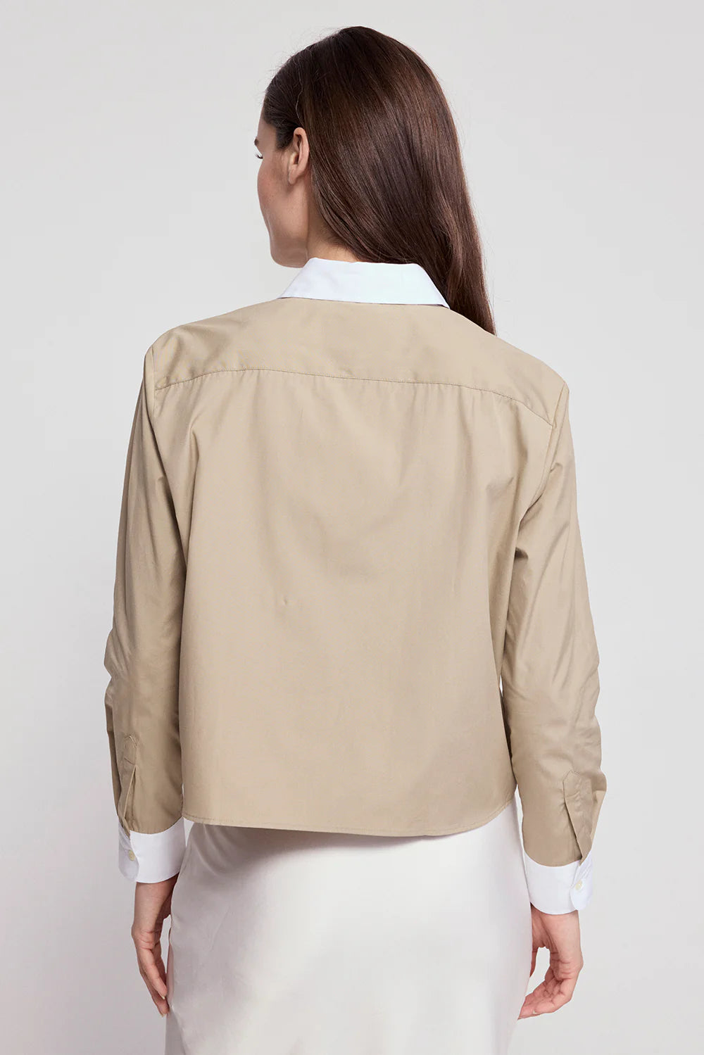 A woman with long brown hair poses with her back to the camera, wearing THE SHIRT&#39;s The LS Boxy Crop beige jacket with white collar and cuffs over a white skirt, against a plain light background.