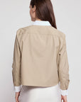 A woman with long brown hair poses with her back to the camera, wearing THE SHIRT's The LS Boxy Crop beige jacket with white collar and cuffs over a white skirt, against a plain light background.