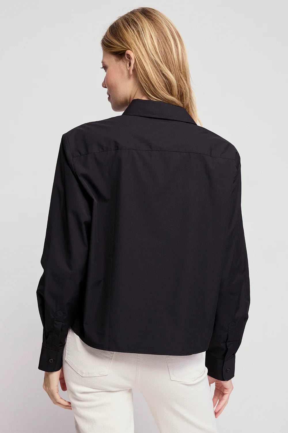 A woman with blonde hair is seen from the back, wearing THE SHIRT’s The LS Boxy Crop—a black, long-sleeve button-up—paired with off-white pants against a plain light grey background.