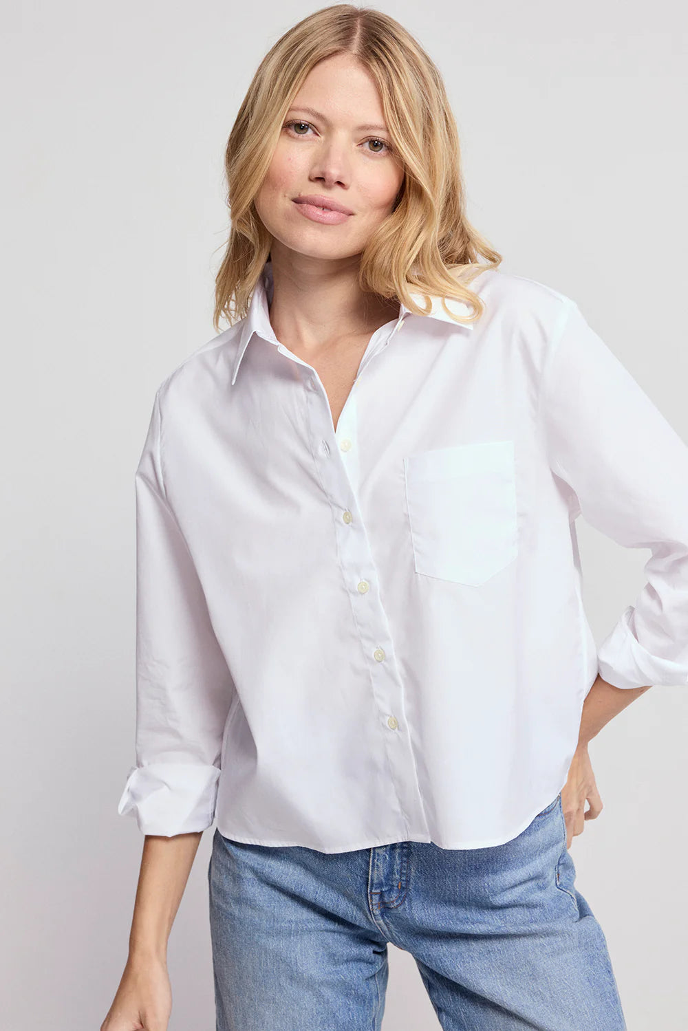 A woman with blonde hair wears THE SHIRT’s The LS Boxy Crop, a classic white cropped shirt with rolled-up sleeves, paired with blue jeans. She stands against a light background and smiles softly at the camera.