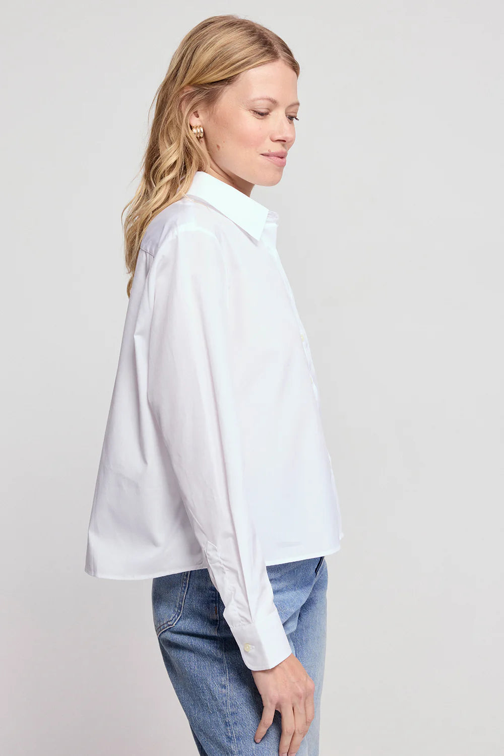 A woman with blonde hair wears THE SHIRT's LS Boxy Crop, a white women’s button-up, paired with blue jeans, standing in profile against a plain light background.