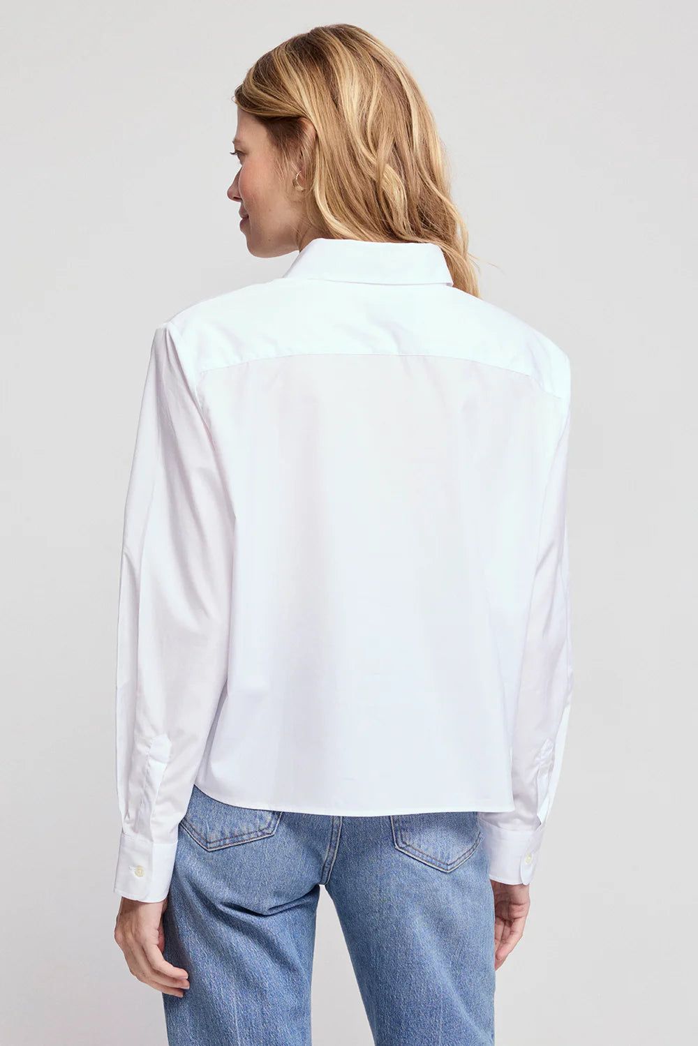A blonde woman wearing THE SHIRT’s The LS Boxy Crop, a long-sleeve white button-up with a No Gape feature, and blue jeans stands facing away from the camera against a light gray background.