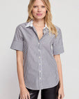 A woman with blonde hair wears THE SHIRT's Short Sleeve Shirt in cotton stretch with black-and-white vertical stripes and No Gape button technology, paired with black pants, against a plain light background, smiling slightly.