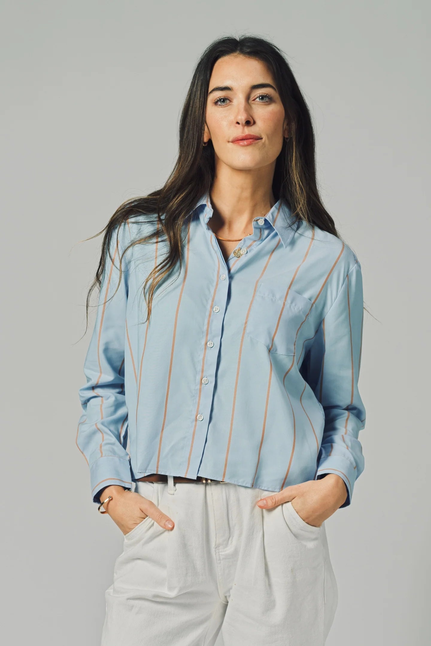 A woman with long dark hair wears THE SHIRT’s The LS Boxy Crop in light blue stripes and white pants, standing with her hands in her pockets against a plain gray background.