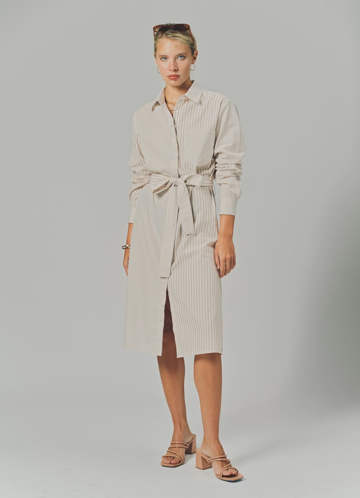 A woman wears THE SHIRT’s LS Midi Dress—a beige and white striped, tie-waist shirtdress—standing against a plain background with her hair tied back, sunglasses on her head, sandals, and a neutral expression.