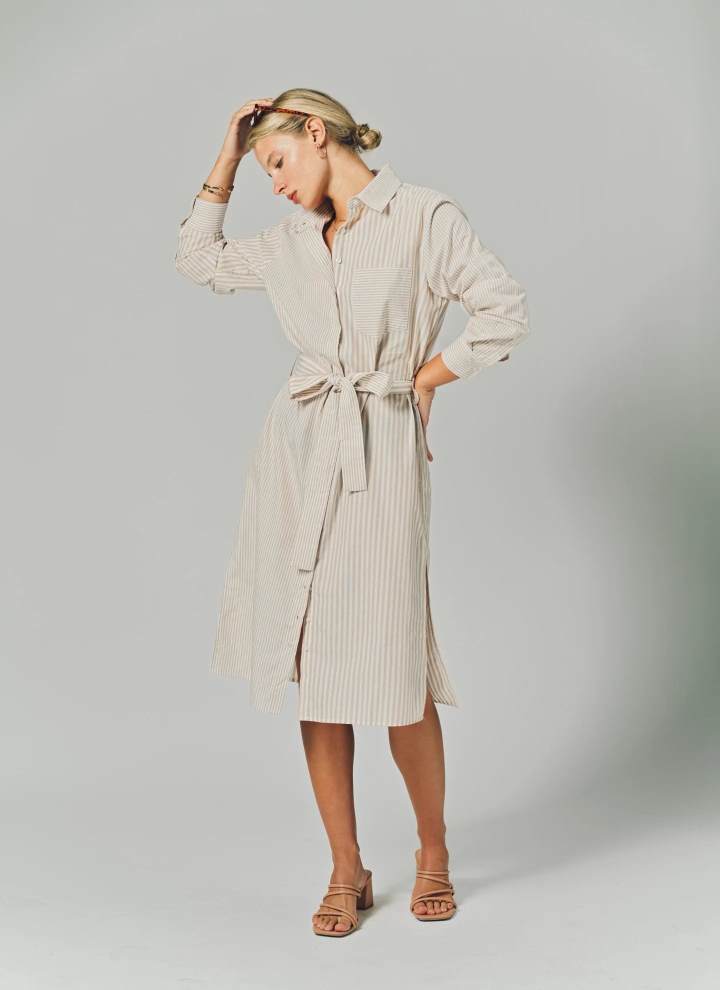 A woman poses in THE SHIRT's LS Midi Dress, a light beige cotton shirtdress with thin vertical stripes, long sleeves, a collared neckline, and a tied waist belt. She completes the look with nude sandals and her hand gently touches her forehead.