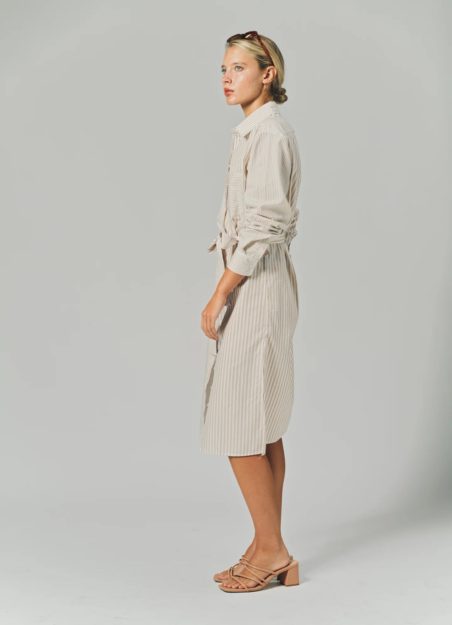 A woman stands in profile against a plain background, wearing THE SHIRT’s LS Midi Dress and tan heeled sandals, with sunglasses perched on her head.