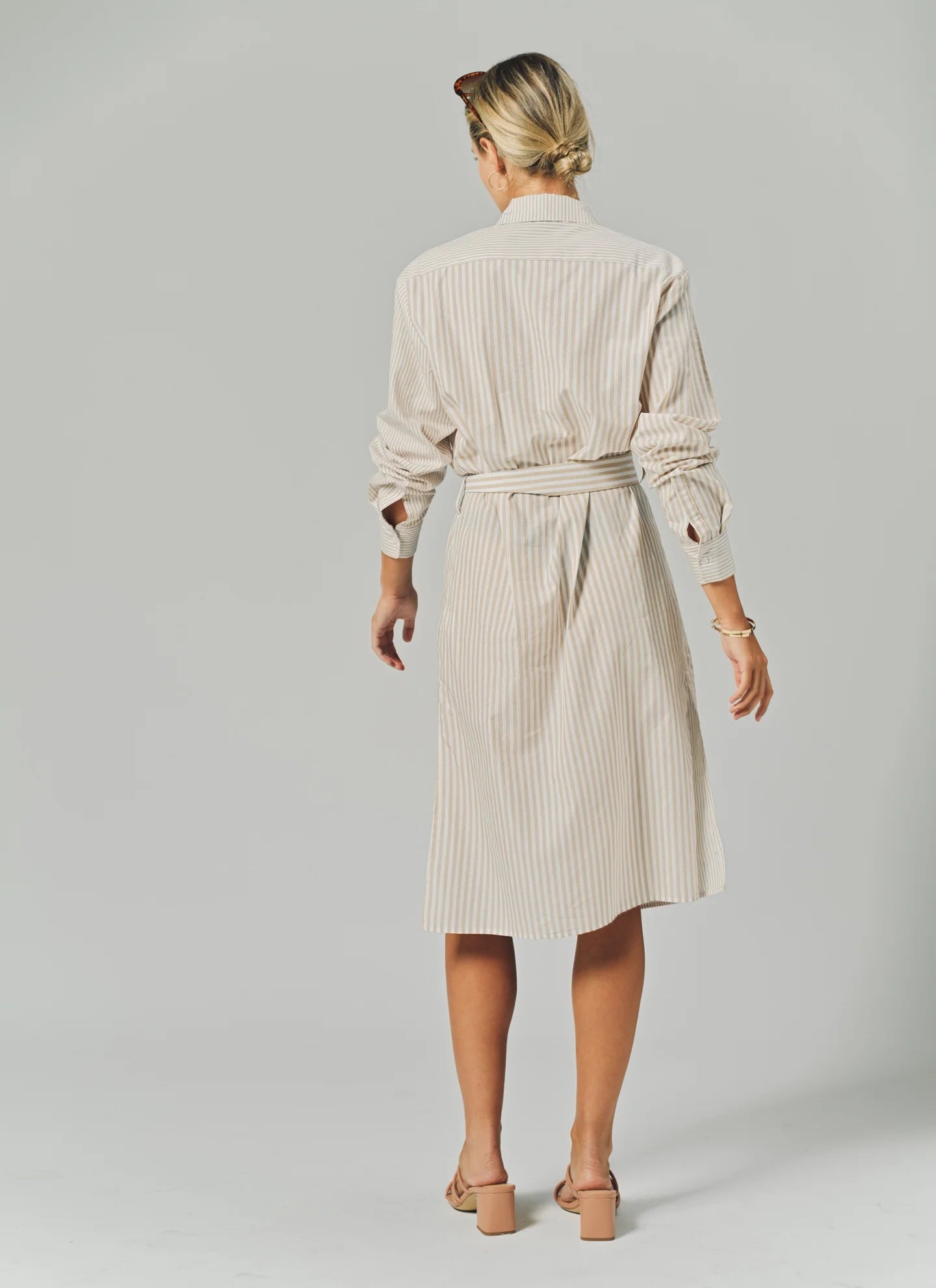 A woman with blonde hair tied back wears THE SHIRT's LS Midi Dress, a knee-length, belted cotton shirtdress with light vertical stripes, paired with light-colored heeled sandals against a plain neutral background.