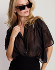 A woman with long blonde hair and black sunglasses wears The Hayden Shirt by THE SHIRT, a sheer lightweight cotton voile blouse, over a black dress against a neutral background, looking slightly to the side.