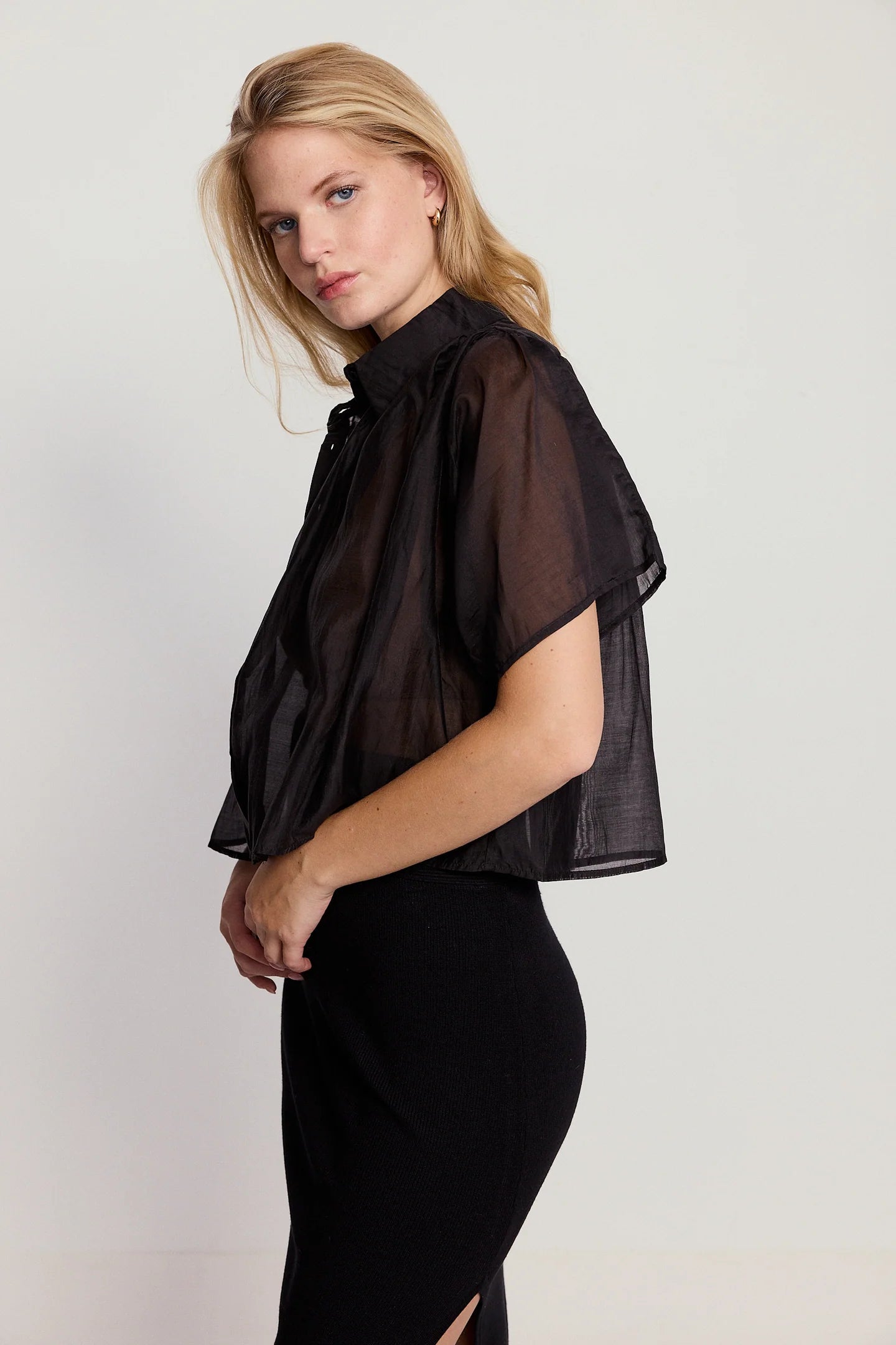 A blonde woman stands sideways against a plain light background, wearing THE SHIRT’s Hayden Shirt in sheer black cotton voile over a black skirt with a side slit. She looks at the camera with a neutral expression.