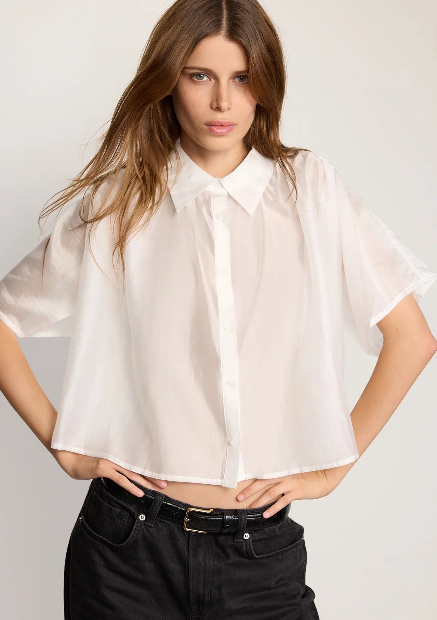 A woman with long brown hair wears THE SHIRT's cropped Hayden Shirt in lightweight cotton voile, paired with black jeans and a belt, standing hands on hips against a plain light background.
