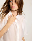 A woman with long brown hair, looking slightly to the side against a plain light background, wears THE SHIRT’s The Hayden Shirt— a short-sleeved, sheer cotton voile style and gently touches her collar.