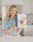 A blonde person in a denim shirt smiles in a bright kitchen, holding the Trader Joe's One Stop Meals Cookbook by Home & Kind, capturing the essence of effortless cooking.