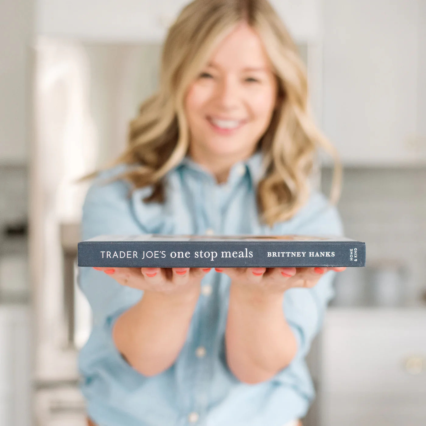 In a softly focused kitchen, a person smiles in a light blue shirt while holding out the "Trader Joe's One Stop Meals Cookbook" by Home & Kind. This book opens up delightful cooking adventures with easy Trader Joe's-inspired recipes.