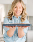 In a softly focused kitchen, a person smiles in a light blue shirt while holding out the "Trader Joe's One Stop Meals Cookbook" by Home & Kind. This book opens up delightful cooking adventures with easy Trader Joe's-inspired recipes.