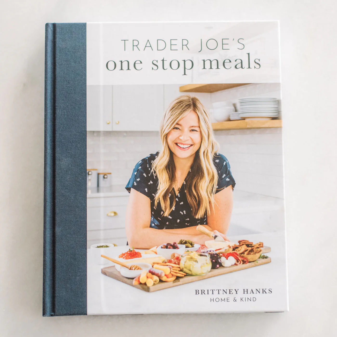 Cover of "Trader Joe's One Stop Meals Cookbook" by Brittney Hanks from Home & Kind. It shows a smiling woman with long blonde hair in a patterned shirt, standing in a kitchen next to a large wooden board filled with Trader Joe's ingredients and easy recipes.