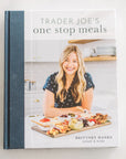 Cover of "Trader Joe's One Stop Meals Cookbook" by Brittney Hanks from Home & Kind. It shows a smiling woman with long blonde hair in a patterned shirt, standing in a kitchen next to a large wooden board filled with Trader Joe's ingredients and easy recipes.