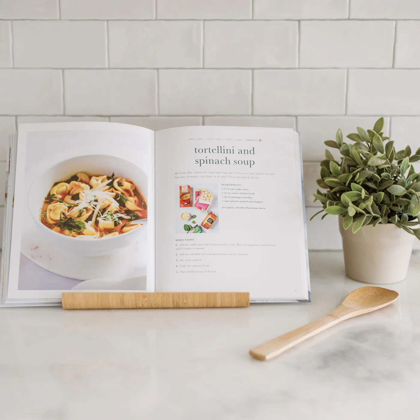 The Trader Joe's One Stop Meals Cookbook by Home & Kind is open on a marble countertop, featuring a tortellini and spinach soup recipe with an image. Nearby, a wooden spoon and potted plant add charm against the tiled backsplash—perfect for exploring new recipes.