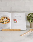 The Trader Joe's One Stop Meals Cookbook by Home & Kind is open on a marble countertop, featuring a tortellini and spinach soup recipe with an image. Nearby, a wooden spoon and potted plant add charm against the tiled backsplash—perfect for exploring new recipes.