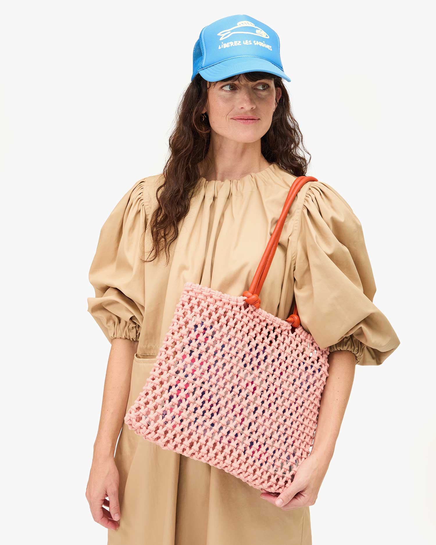 A person with long hair stands against a plain white background, wearing a beige dress and Clare Vivier's Trucker Hat Les Sardines featuring an adjustable snap-back closure. They hold a pink crocheted tote bag with orange handles.