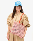 A person with long hair stands against a plain white background, wearing a beige dress and Clare Vivier's Trucker Hat Les Sardines featuring an adjustable snap-back closure. They hold a pink crocheted tote bag with orange handles.