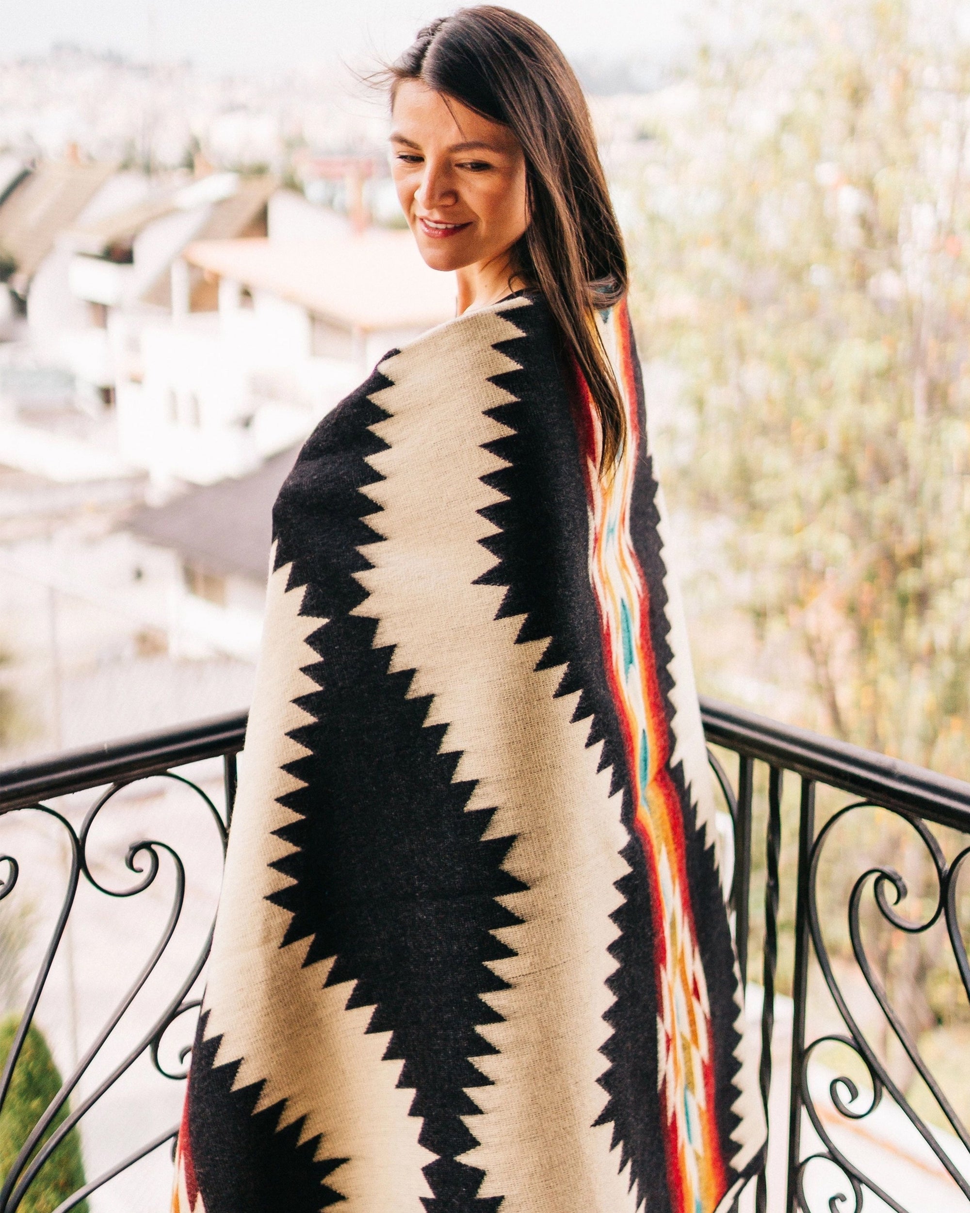 A woman smiles on a balcony, wrapped in the Faire Tsachila Blanket featuring bold zigzag and colorful geometric patterns. Blurred houses and trees are in the background.