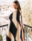A woman smiles on a balcony, wrapped in the Faire Tsachila Blanket featuring bold zigzag and colorful geometric patterns. Blurred houses and trees are in the background.