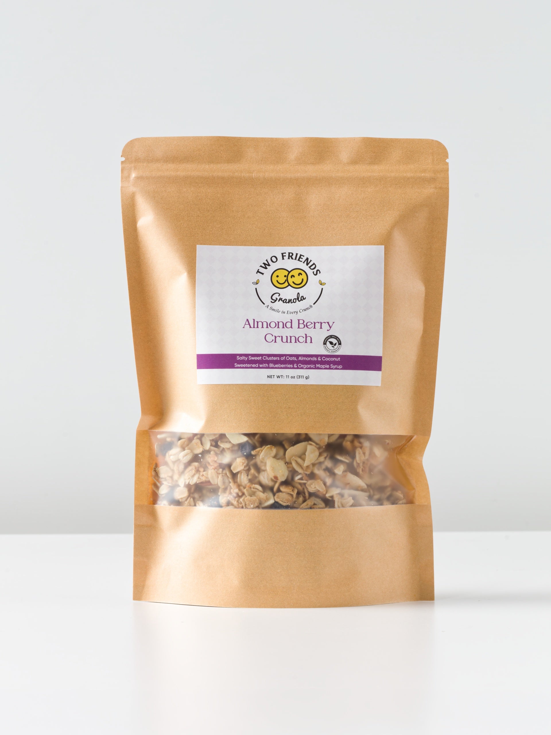 A brown resealable pouch of Two Friends Granola Almond Berry Crunch sits on a white surface, featuring crunchy almonds, a clear window displaying granola inside, and a white label with a purple stripe.