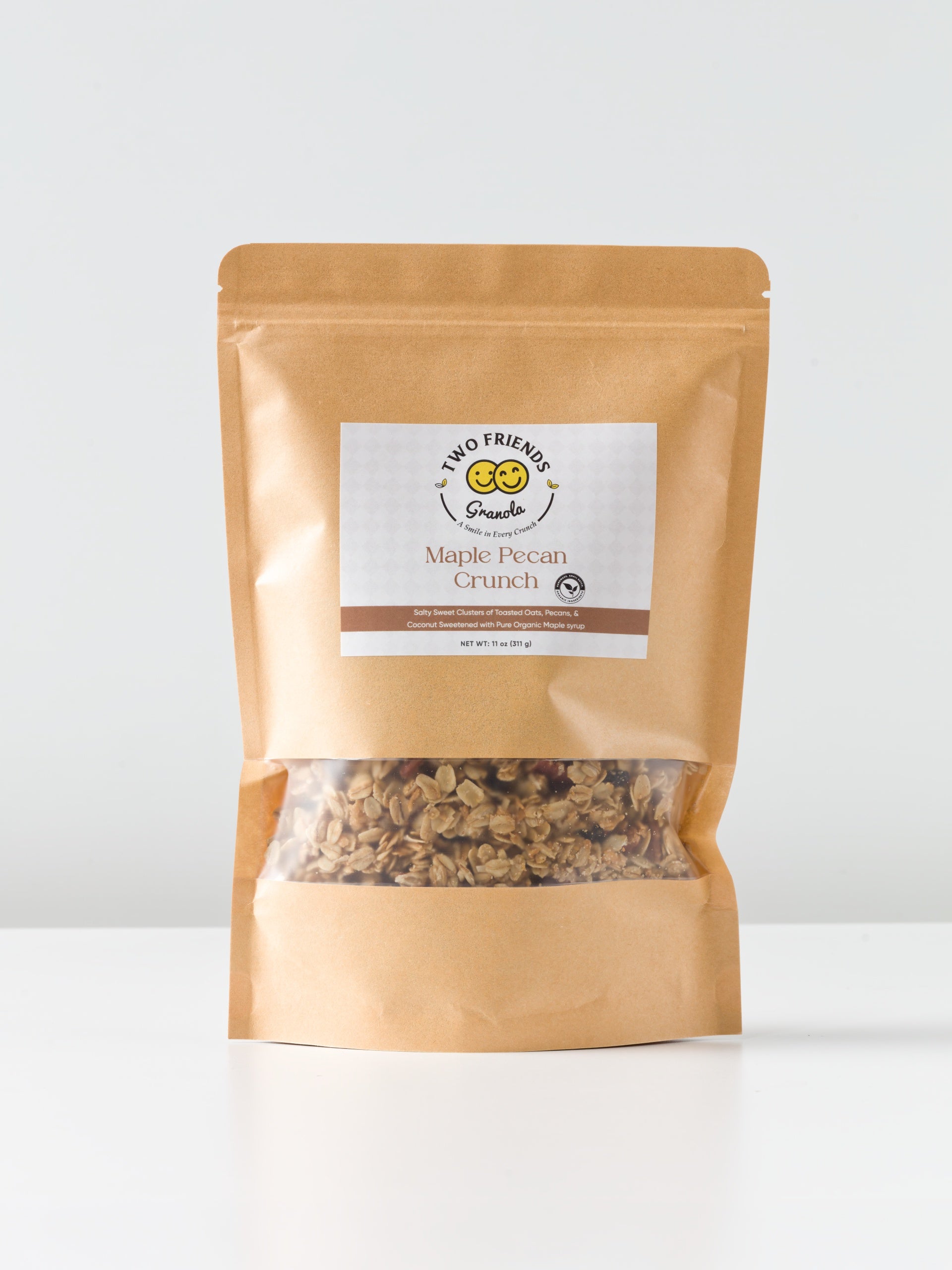 A brown resealable pouch labeled "Two Friends Granola Maple Nut Crunch" sits on a white surface, with golden oats and granola visible through a clear window at the bottom of the package.