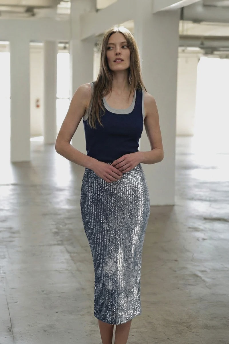 A woman with straight light brown hair poses indoors, wearing the Le Superbe Liza Sequin Skirt and a navy tank top, her hands clasped in a spacious, sunlit room.