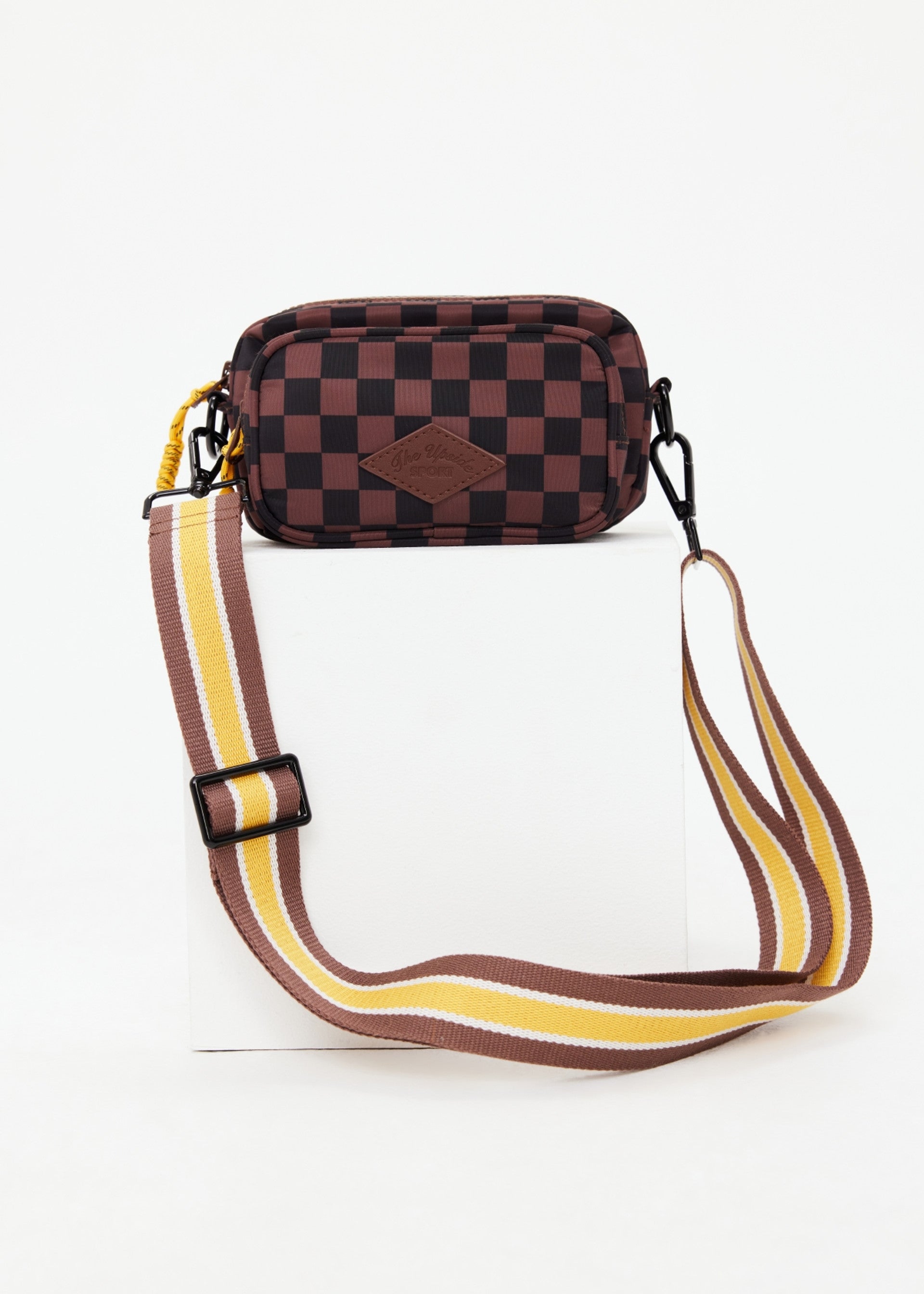 The Upside TECHNICA VOYAGE CROSSBODY BAG features a dark brown and black checkered pattern, front leather patch, and a wide adjustable strap in brown, yellow, and white. Made from recycled polyester and displayed on a white block.