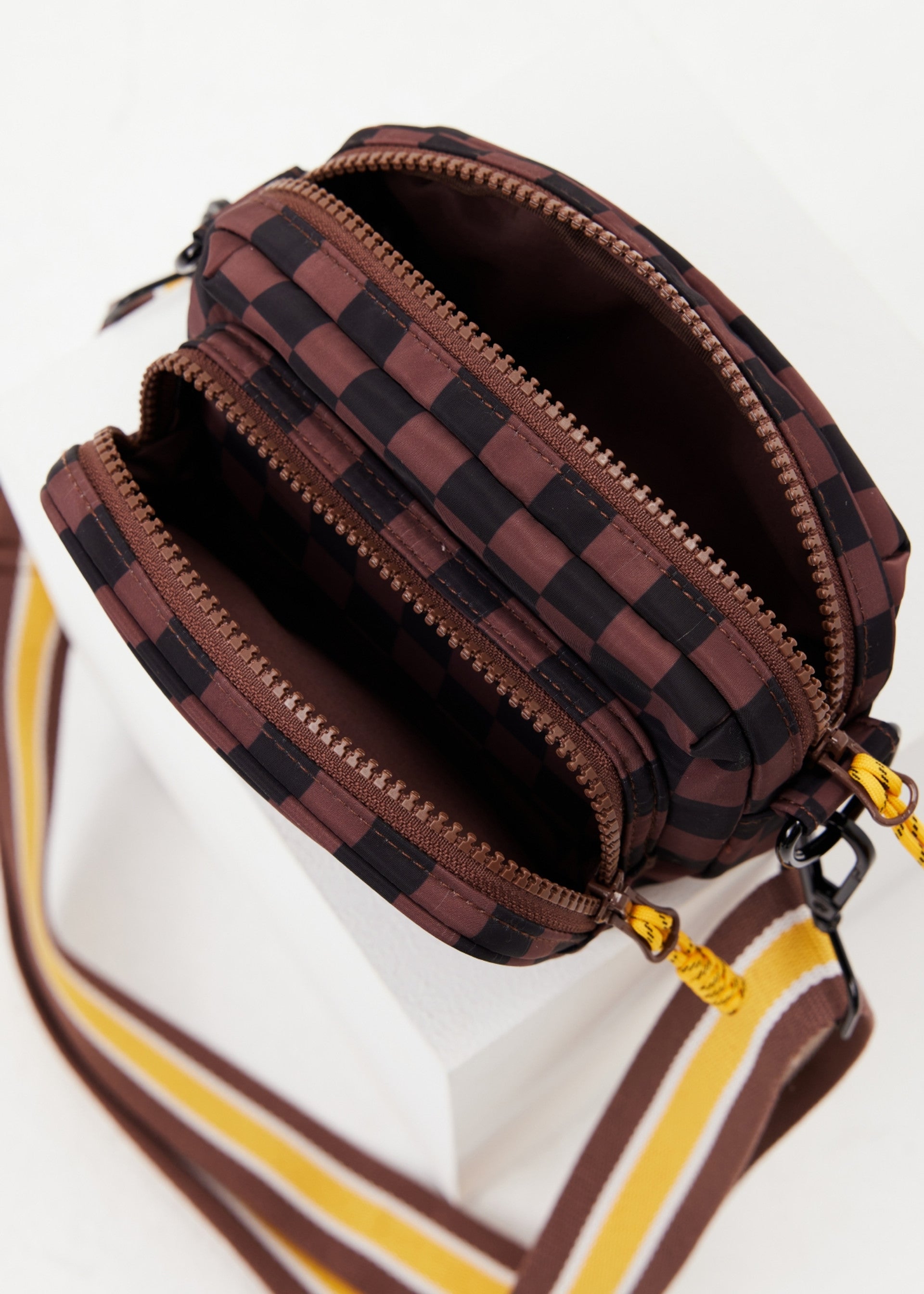 The TECHNICA VOYAGE CROSSBODY BAG by The Upside is a small rectangular bag with a brown and black checkered design, two zippered compartments, and a wide adjustable strap in brown, yellow, and white for comfort.