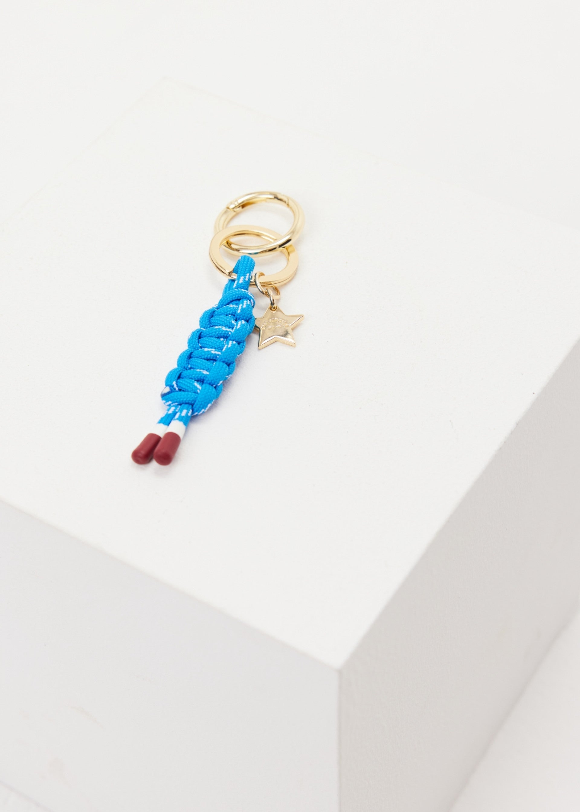 The Upside LIMITLESS KEYRING features a gold charm with braided blue macrame paracord, red-tipped ends, and a small gold star, shown on a white geometric surface.