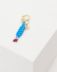 The Upside LIMITLESS KEYRING features a gold charm with braided blue macrame paracord, red-tipped ends, and a small gold star, shown on a white geometric surface.