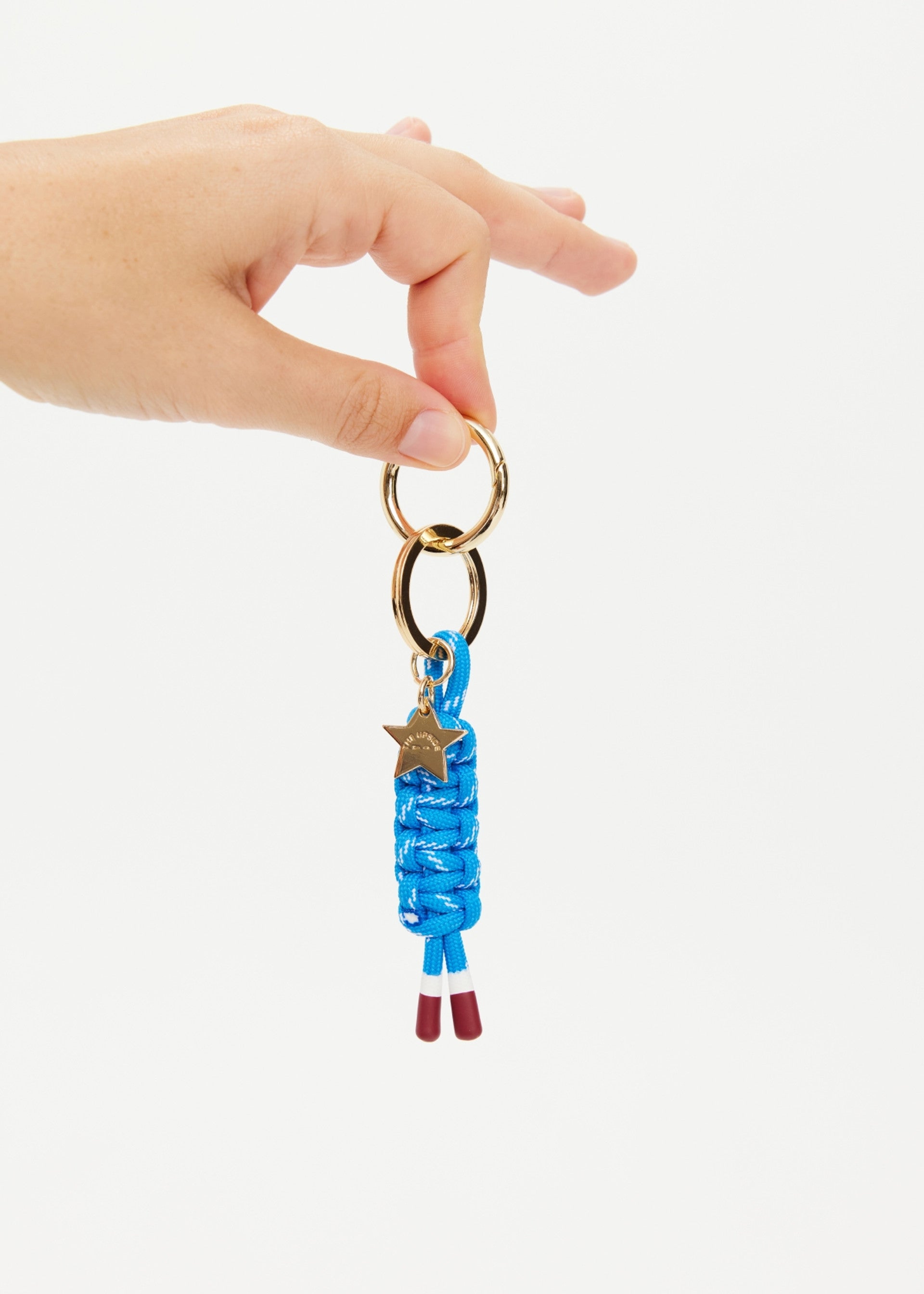 A hand holds The Upside LIMITLESS KEYRING, featuring gold metal hardware, a blue macrame paracord with a gold star charm, and two brown-tipped cord ends, shown against a plain white background.